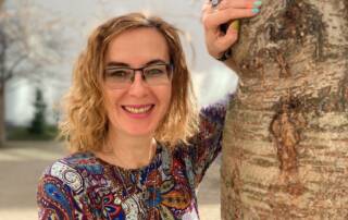 Victoria Aharonov hat lange dunkelblonde Haare, trägt eine Brille und eine gemusterte Bluse